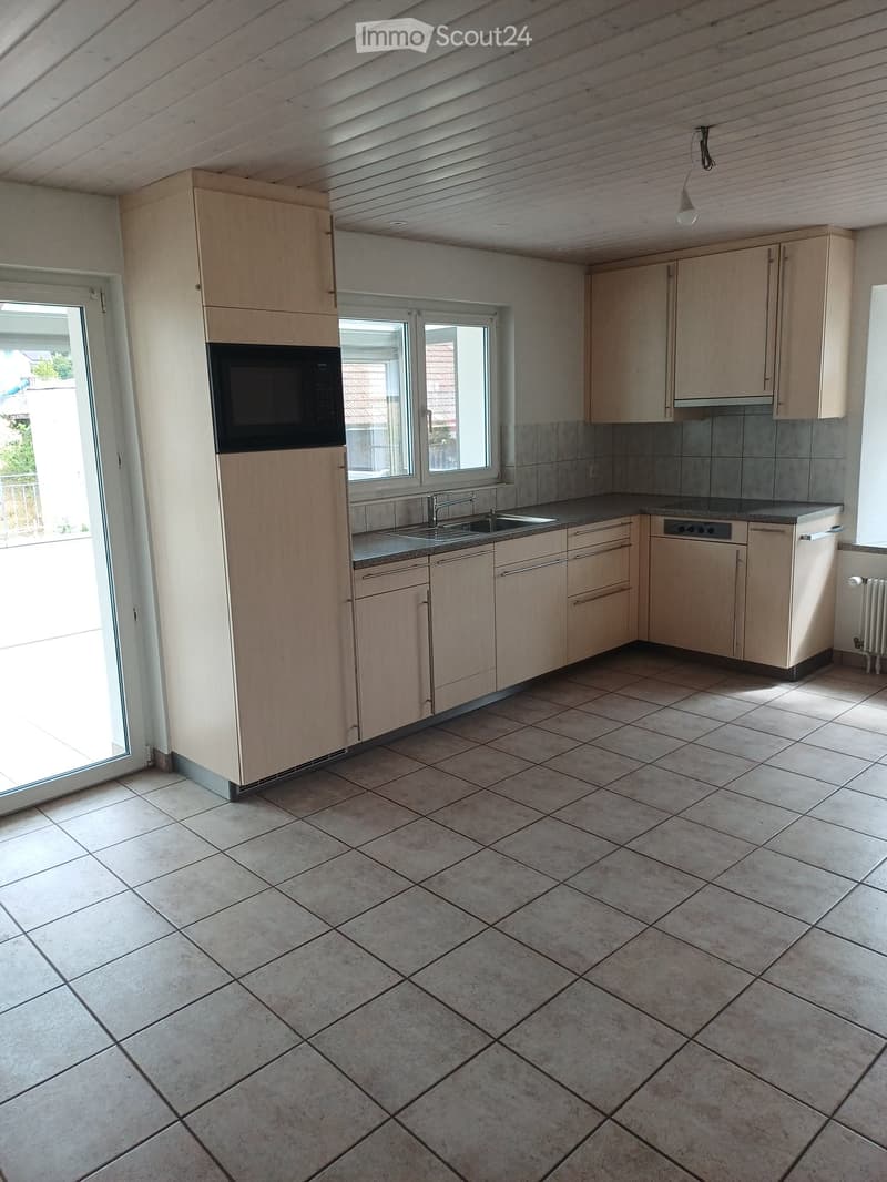 Modern kitchen with light wooden cabinets, tiled floor, microwave, sink, and large windows letting in natural light.