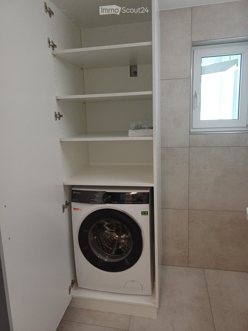 A compact bathroom with a built-in washing machine in a cabinet, white tiled walls, and a window providing natural light.