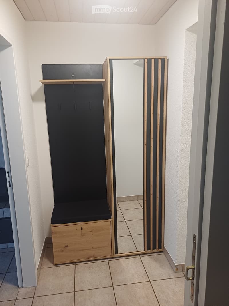 Hallway with tiled floor, white walls, a wooden bench with a black cushion, and a mirror with wooden frame.