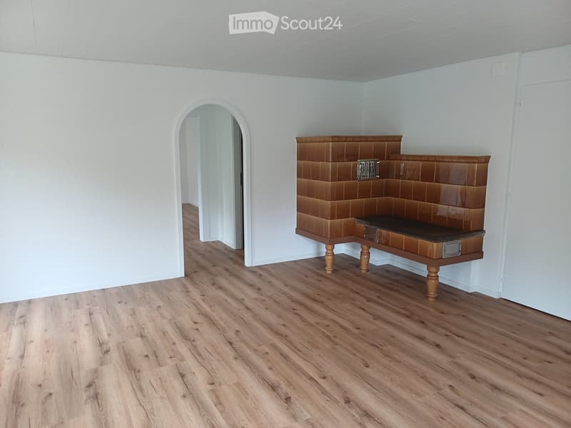 A small, unfurnished room with light wooden flooring and white walls. A built-in bench with a dark surface and wooden legs is positioned against one wall. An arched doorway leads to another space.