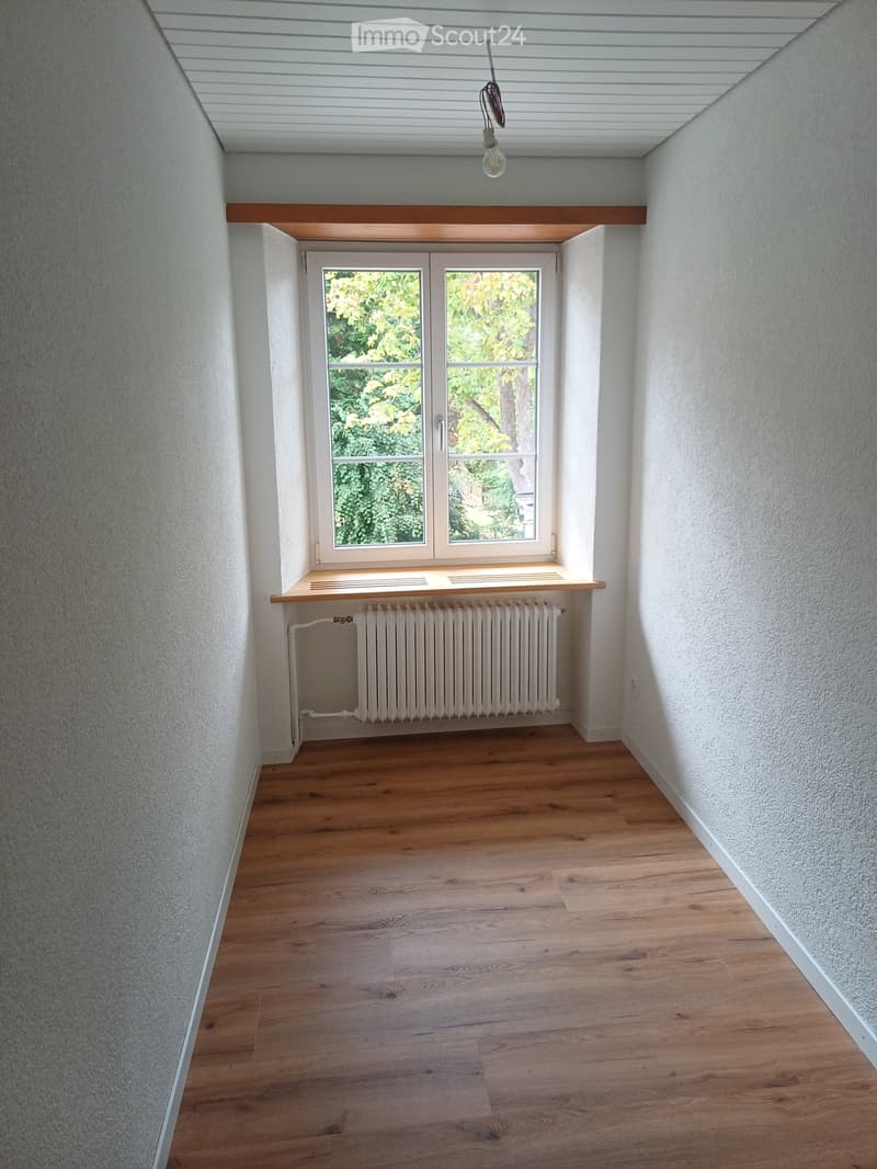 Empty room with wooden flooring, white walls, a window with a view of greenery, a radiator, and a small wooden shelf above it.