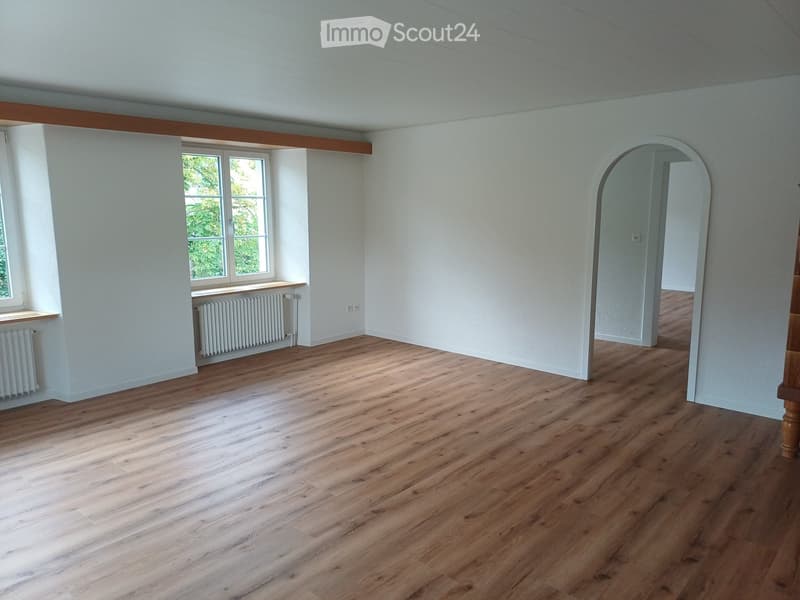 Empty room with light wooden flooring, white walls, and large windows allowing natural light. Radiators are visible beneath the windows.