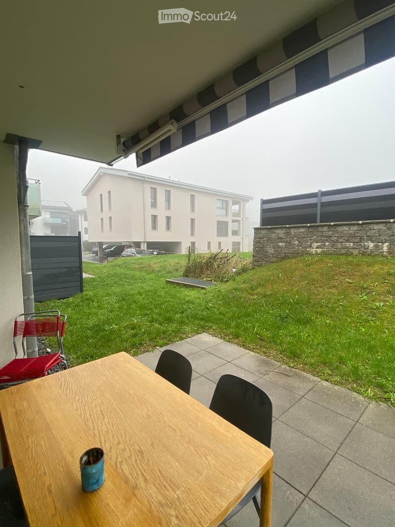 Outdoor patio area with a wooden table, chairs, and a cup, overlooking a grassy yard with a modern building in the background.
