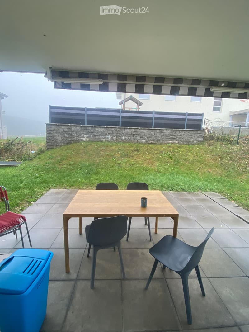 Outdoor patio area with a wooden table, four chairs, and a cup on the table. The patio is covered by an awning and has a stone border and grassy area.