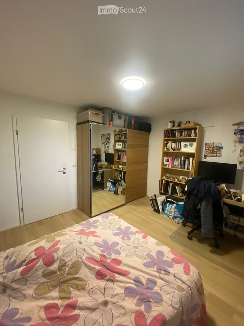 A well-lit bedroom with a wooden floor, a bed with a floral duvet, a wardrobe, a bookshelf, a chair, and a screen.