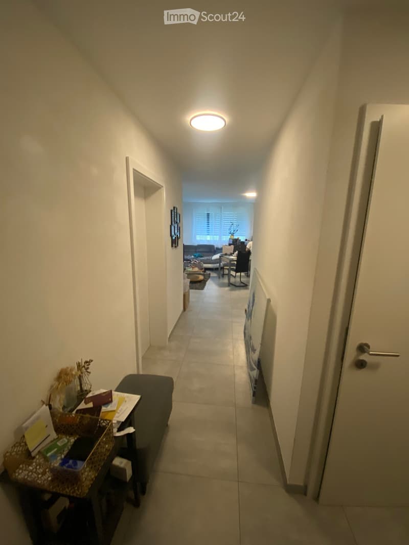 A narrow hallway with tiled flooring, white walls, and a ceiling light. A small table with items sits on the left, and a door is visible on the right. The hallway leads to an open area with furniture visible in the background.