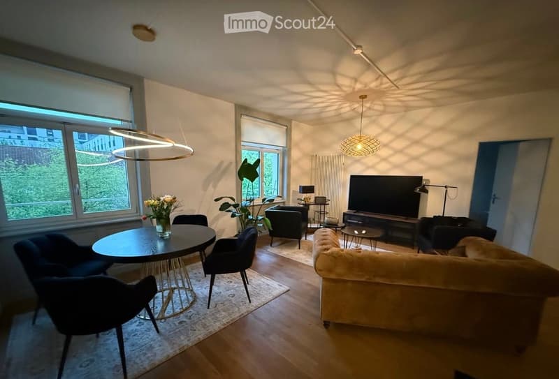 Modern living room with a cream-colored sofa, coffee table, dining table with chairs, and a television. The room features large windows with a view of greenery, pendant lights, and a small side table with a lamp.