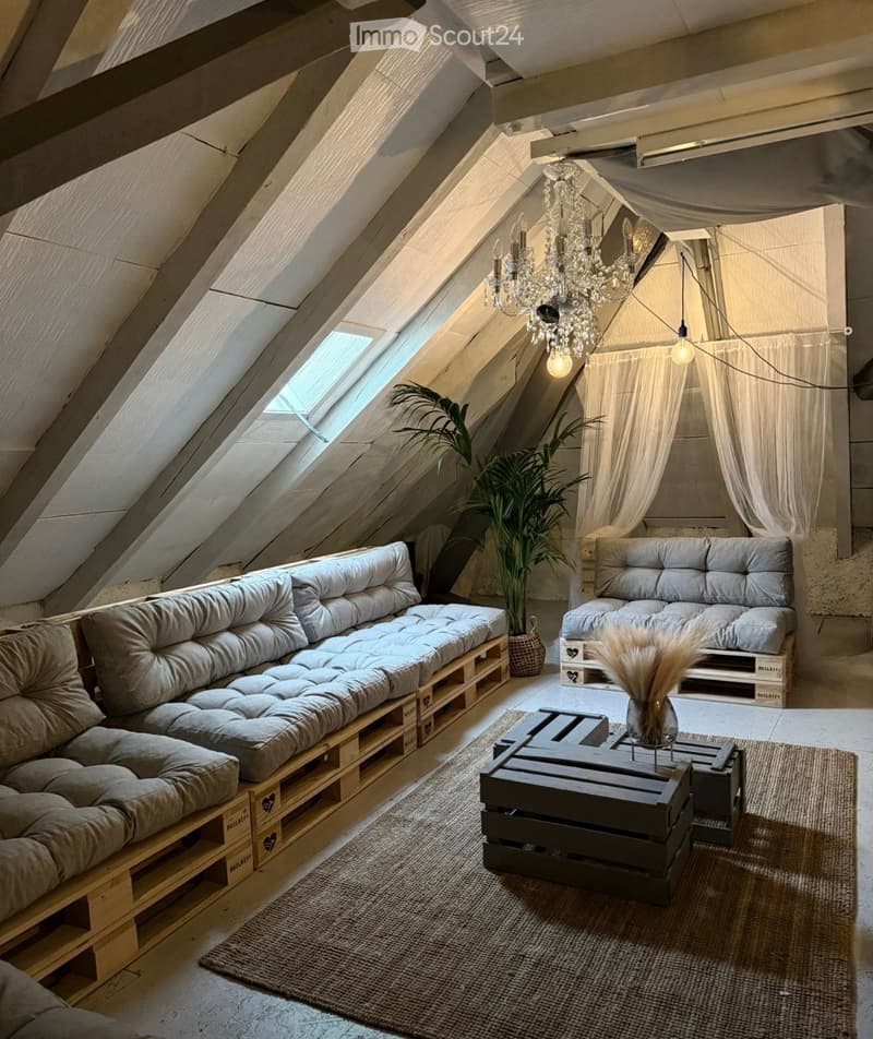 Cozy attic living space with slanted wooden ceilings, skylight, grey sofa, coffee table, chandelier, and plants.