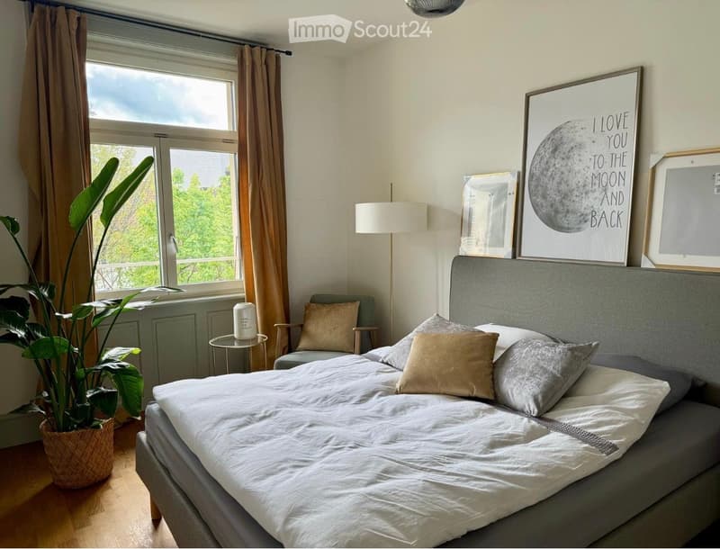 A modern, well-lit bedroom with a large bed, pillows, a grey headboard, a small round table with a plant, a chair, and a floor lamp. The room features large windows with curtains and several picture frames on the wall.