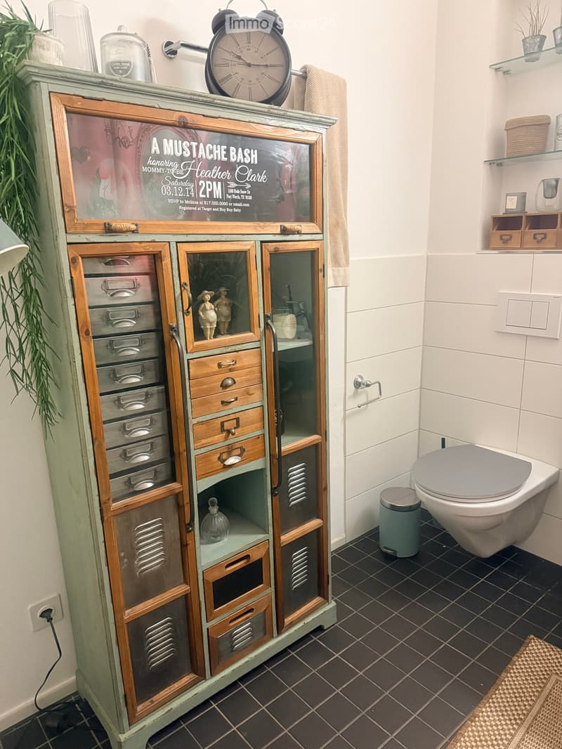 Bathroom with a vintage-style wooden cabinet, a toilet, a wall-mounted clock, and a trash bin. The cabinet has multiple drawers and glass-fronted compartments, and the walls are tiled with white tiles.