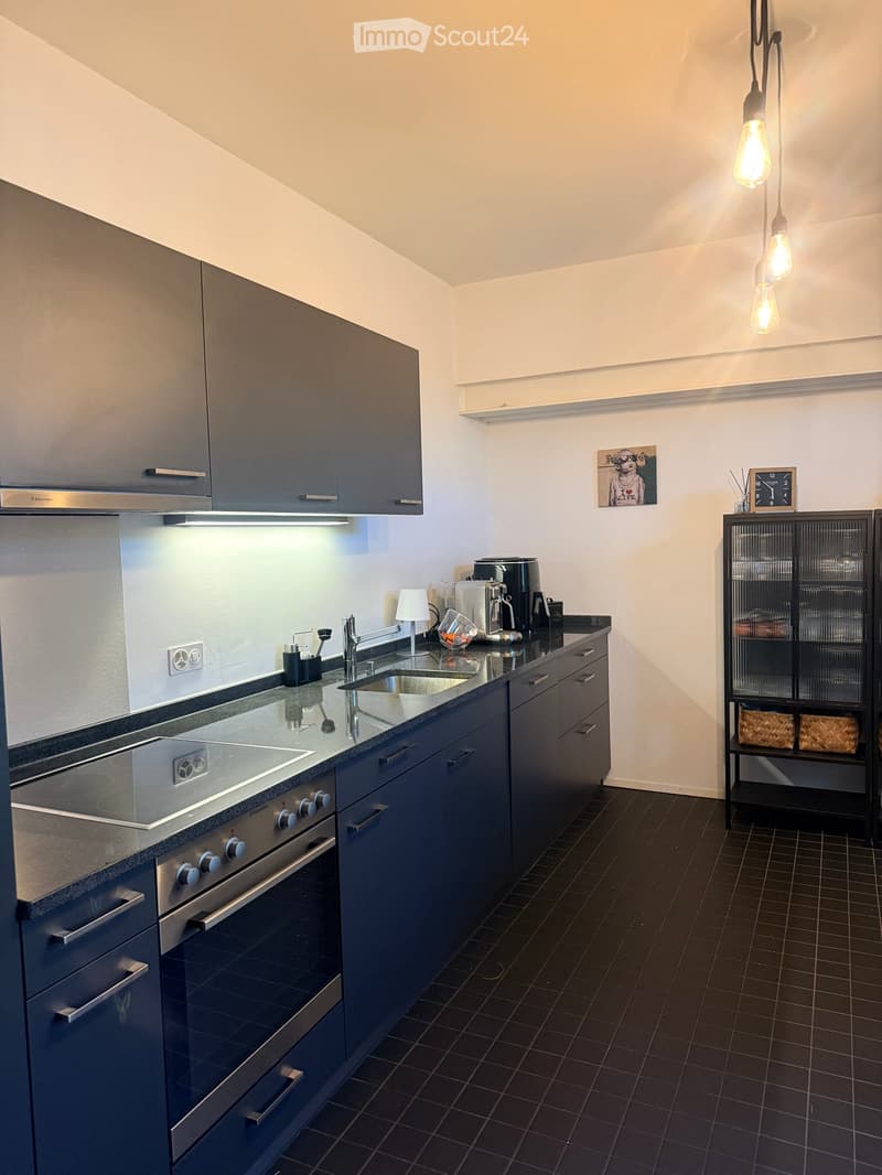 Modern kitchen with dark blue cabinets, stainless steel appliances, and a black tiled floor. Features include a sink, oven, coffee maker, and a small shelf with decorative items.