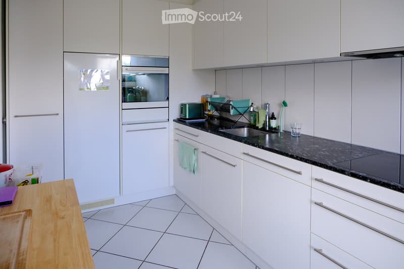 Modern kitchen with white cabinets, black granite countertops, stainless steel sink, and various appliances including a refrigerator, oven, and toaster. The space features a wooden cutting board and a dish rack with cups and bottles.