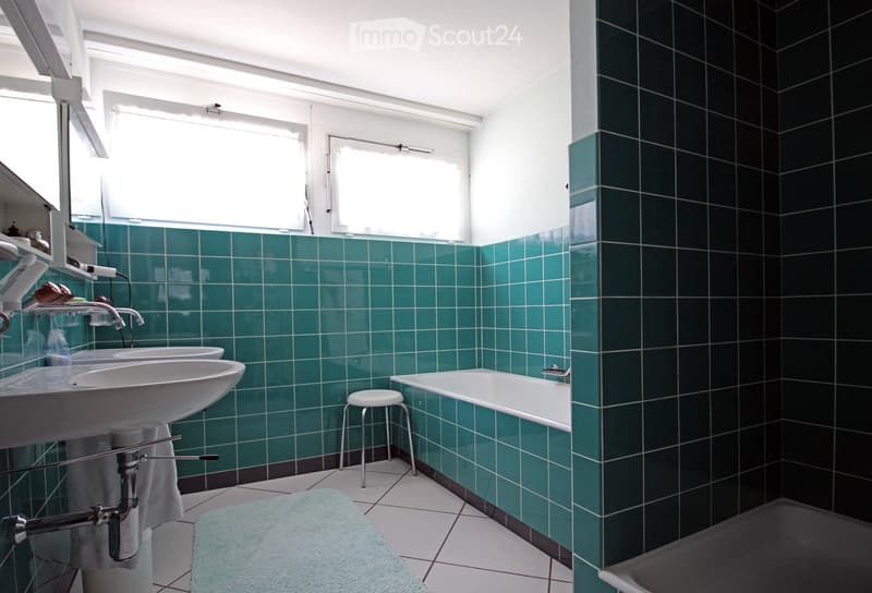 Bathroom with green tiled walls, white tiled floor, double sink, bathtub, small stool, and a mirror above the sink.