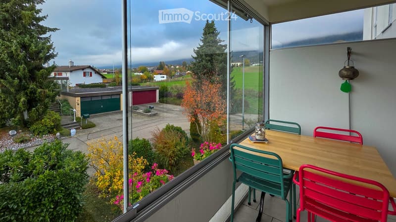 Balcony with wooden table and colorful chairs, overlooking a landscaped garden and residential area.