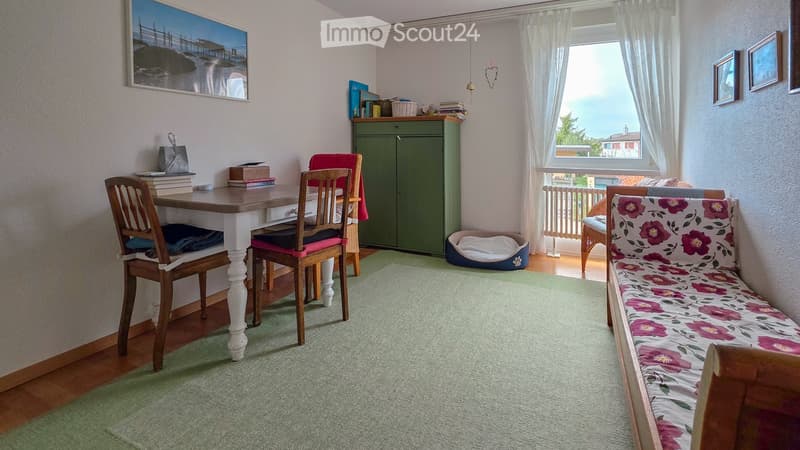 A cozy, well-lit room with a wooden table and chairs, a floral-patterned sofa, a green cabinet, and a pet bed. The room features a large window with white curtains and a view of the outdoors.