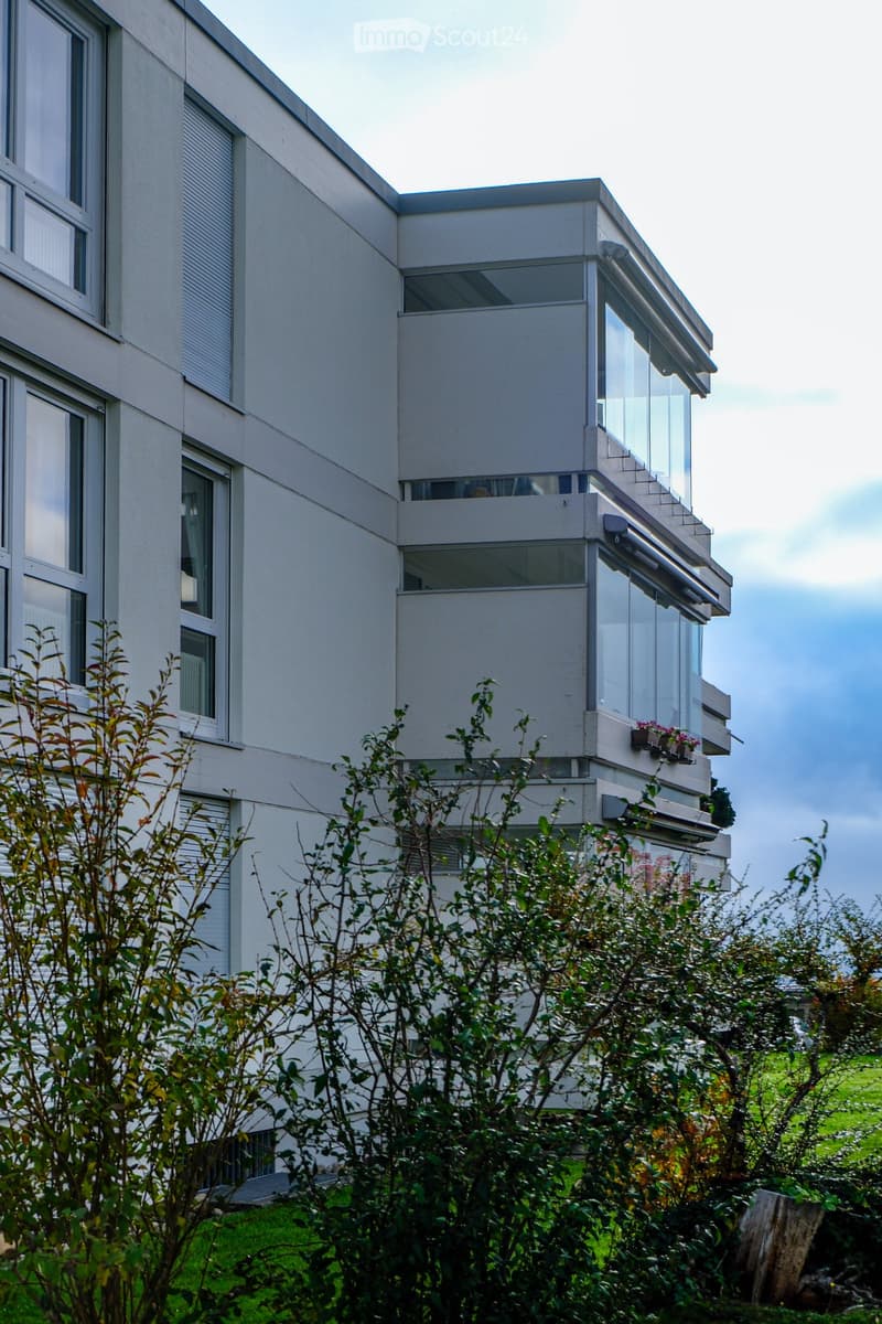 Modern multi-story building with large windows and balconies, surrounded by greenery.