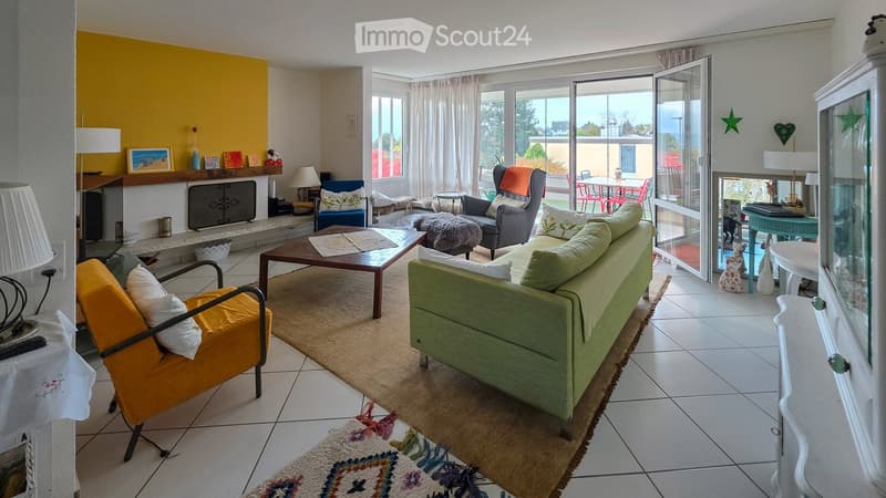 Bright living room with large windows and sliding glass doors leading to a patio. Features a green sofa, wooden coffee table, colorful armchairs, and a cozy rug.