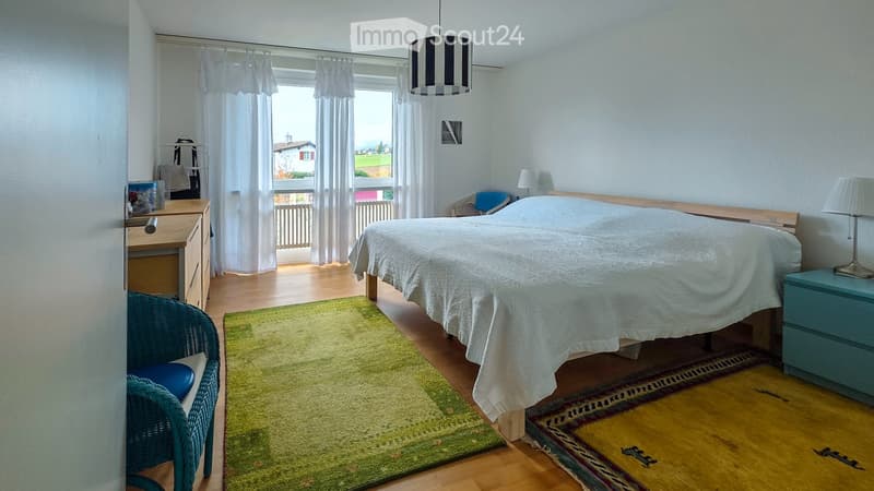 Bright bedroom with a large bed, nightstand, desk, chair, and large windows with curtains. The room features wooden flooring and a green rug.