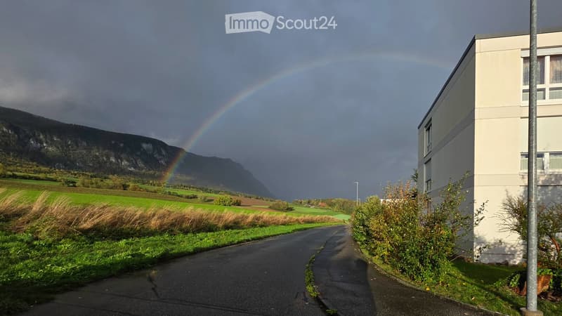 Modern building with white facade and multiple windows, situated on a road with a rainbow visible in the background and a mountain range in the distance.