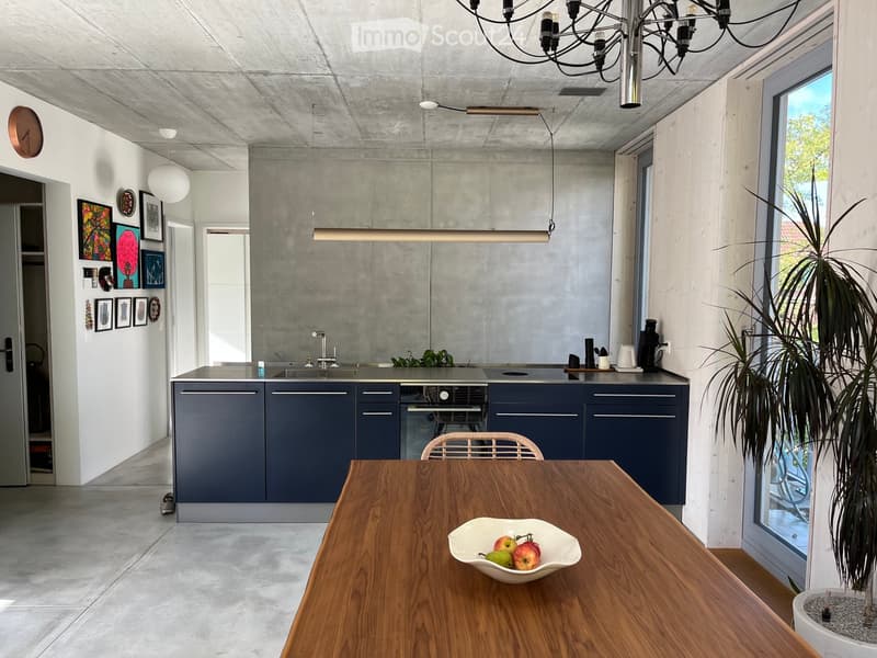 Modern kitchen with dark blue cabinets, wooden dining table, bowl of fruit, pendant lights, and framed art on the wall.