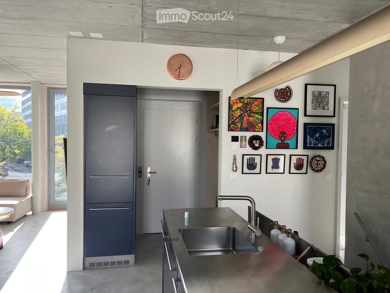 Modern kitchen with stainless steel sink, faucet, and countertop. Features framed artwork on the wall, a clock, and a small sofa visible in the adjacent space.