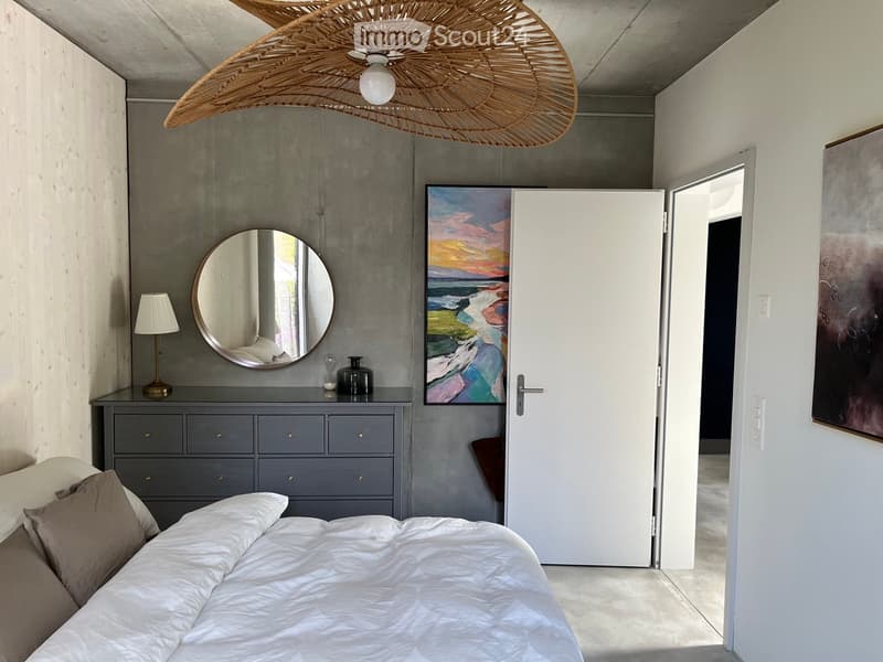 Modern bedroom with a large bed, nightstand, lamp, mirror, and artwork on the walls. Features a wicker hanging light fixture and a grey dresser with decorative items.