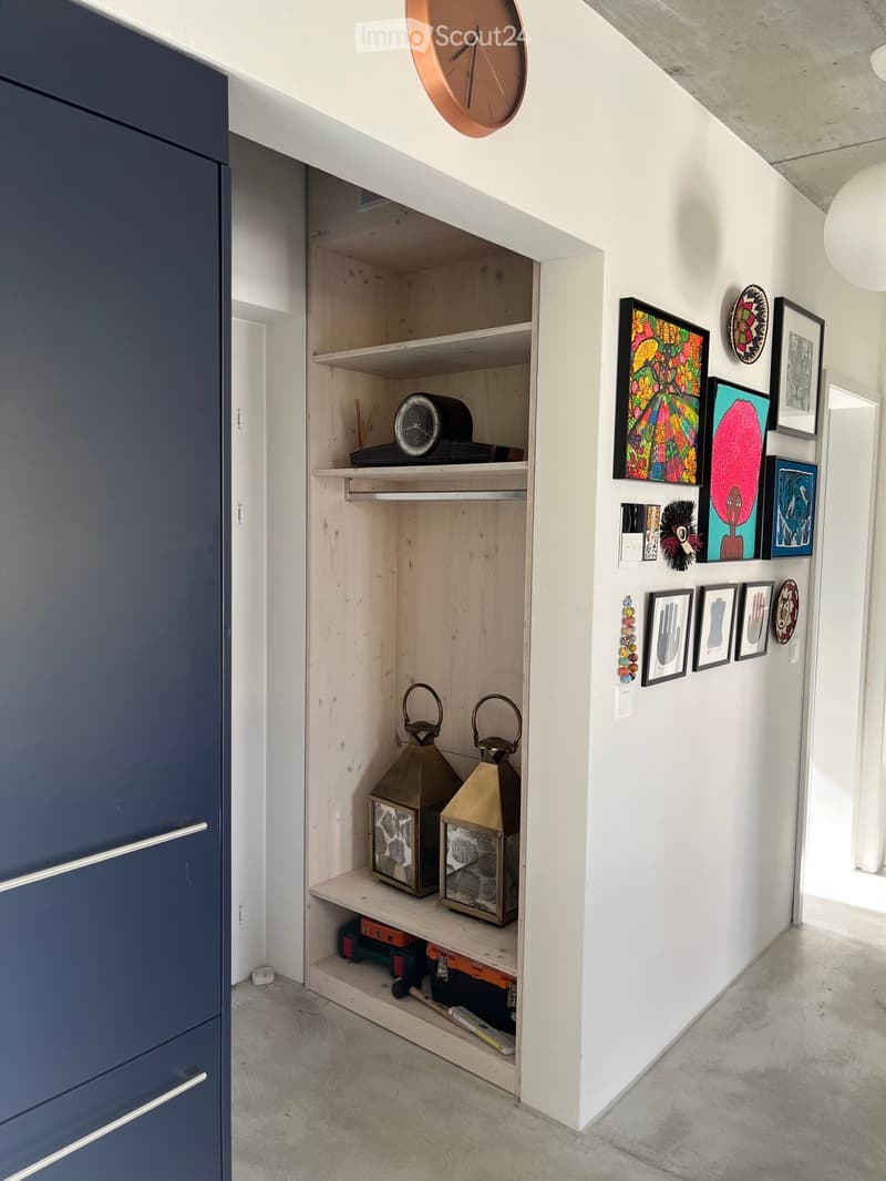 Modern hallway with white walls, concrete floor, built-in wooden shelving unit, and various decorative items including framed artwork and lanterns.