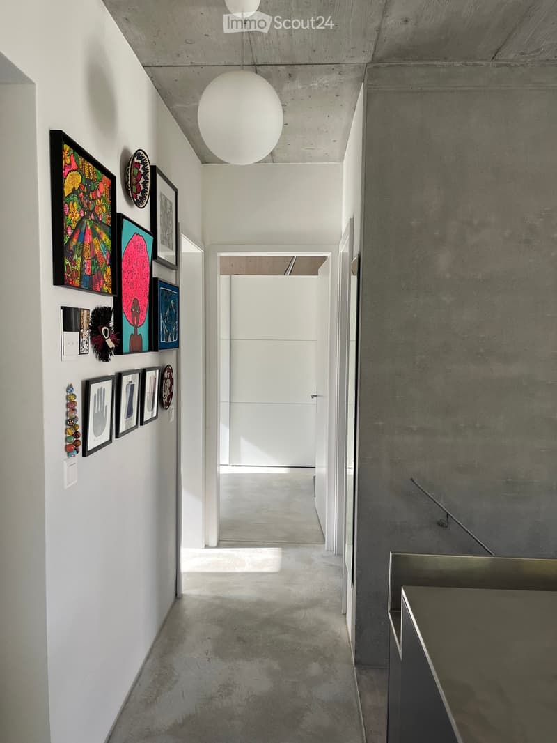 Modern hallway with white walls, concrete floor, and a large spherical pendant light. Various framed artworks and decorative plates are displayed on the left wall, leading to an open doorway at the end.