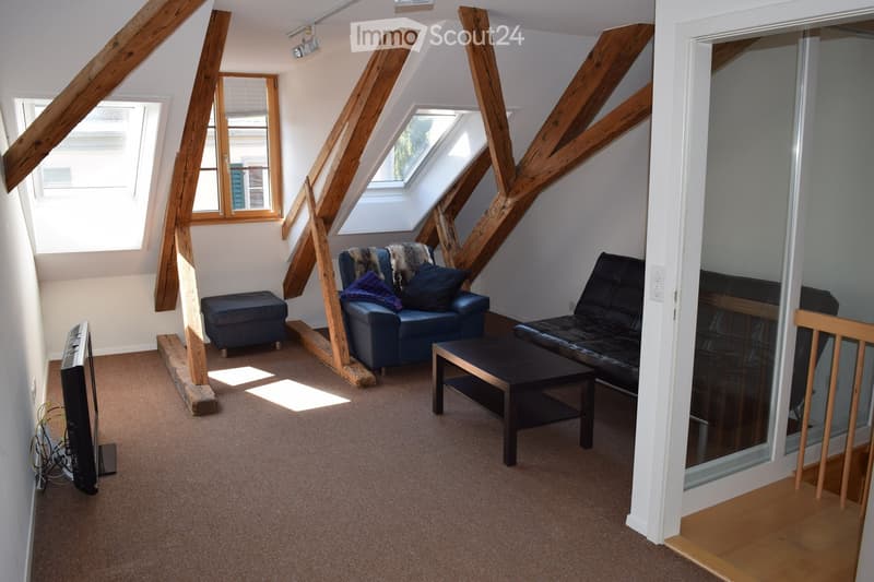 Cozy attic living room with exposed wooden beams, large windows, and skylights. Features a dark blue sofa, black leather chair, coffee table, ottoman, and television.