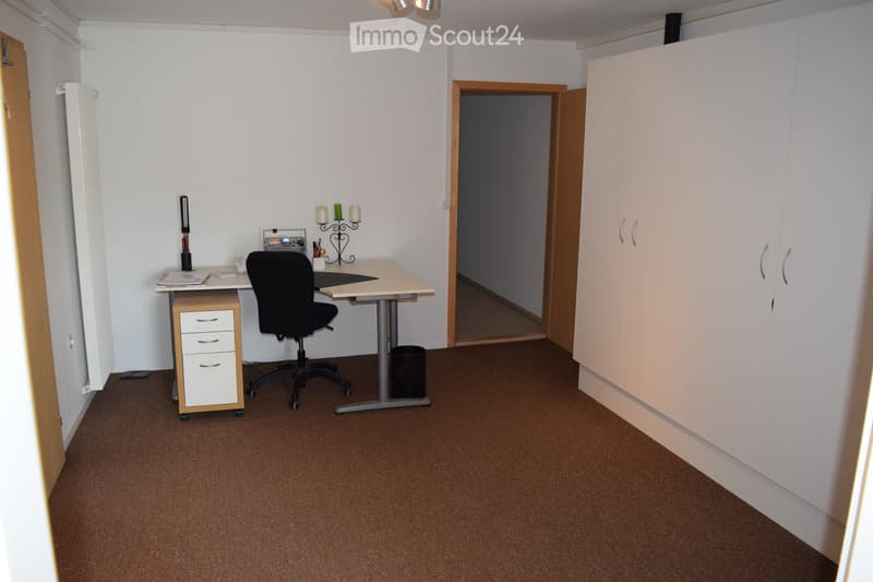 A furnished room with a desk, chair, cabinet, and candle holder. The room has white walls, brown carpeting, and a door leading to another space.