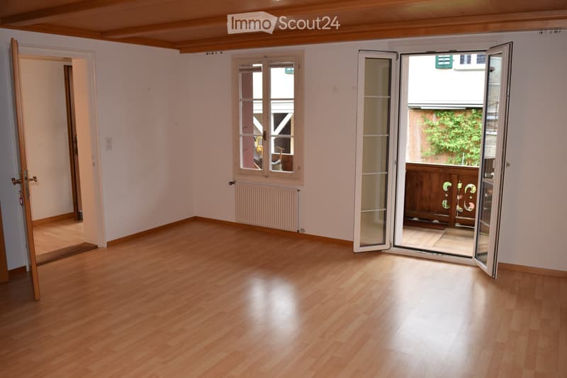 Empty room with wooden flooring, white walls, large windows, and an open sliding door leading to a balcony or outdoor area.