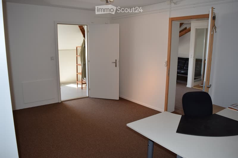Office space with a white table, black chair, and a coat rack in the adjacent room.