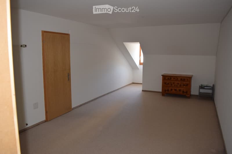 A small, carpeted room with white walls and a slanted ceiling. Features include a wooden door, a window with a skylight, a chest of drawers, and a small table with books.