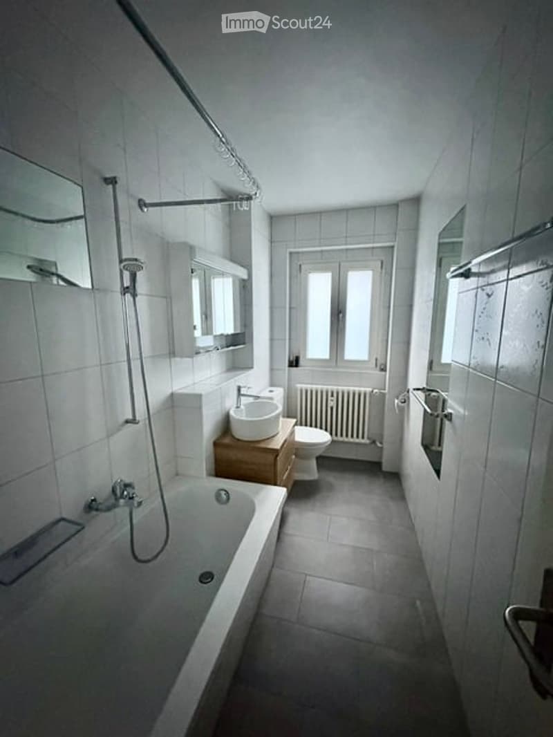 Modern bathroom with white tiles, a bathtub, toilet, sink, and shower. Features include a large mirror, a radiator, and a window for natural light.