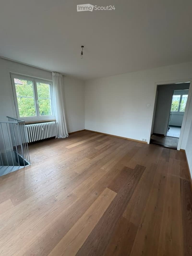 Empty room with wooden flooring, white walls, large window with white curtains, radiator, and a staircase leading upwards.