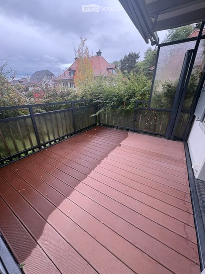 Wooden deck with black railing, overlooking a residential area with trees and houses.