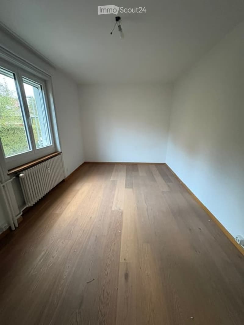 Empty room with wooden flooring, white walls, and a single window with a radiator below it.