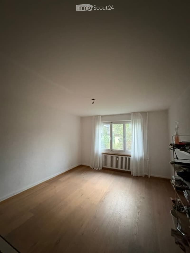 Empty room with wooden flooring, white walls, a window with white curtains, and a radiator below the window. No furniture or equipment is present.