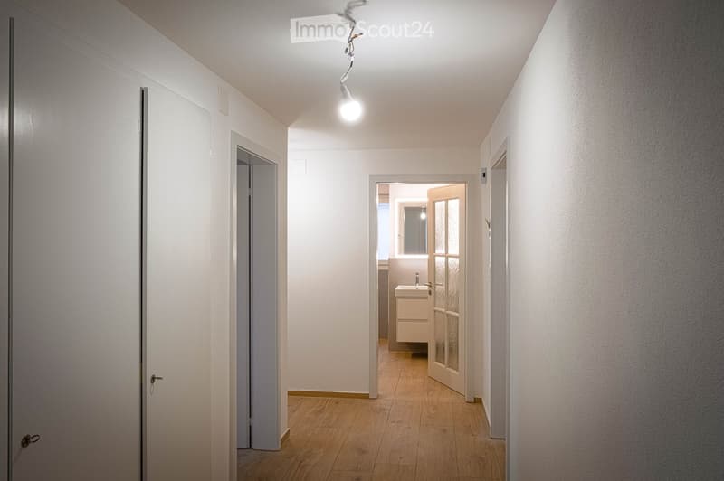 A clean, well-lit hallway with wooden flooring, white walls, and several doors leading to different rooms, including a visible bathroom with a sink.