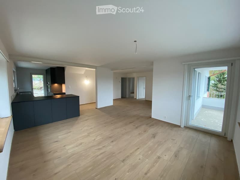 Empty room with wooden flooring, white walls, and a dark kitchen counter visible in the background. A sliding glass door leads to a balcony area.