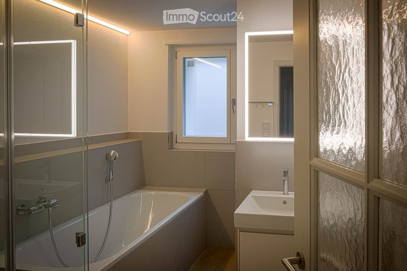 Modern bathroom with a bathtub, sink, and shower area. Features include a large window, built-in lighting, and sleek fixtures.
