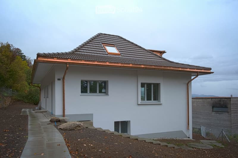 Single-story house with white exterior, grey tiled roof, copper gutters, and multiple windows. The house features a raised ground floor with a concrete pathway leading to the entrance.