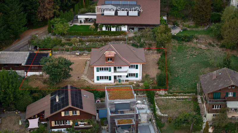 Aerial view of a residential area with multiple houses, including a prominent white house with green shutters and a brown roof, surrounded by greenery and neighboring homes.