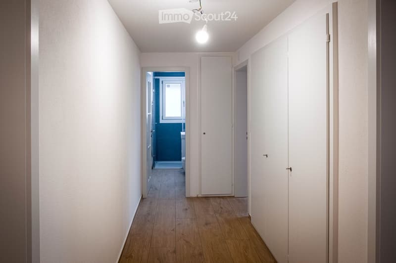 A clean, well-lit hallway with wooden flooring and white walls, featuring several closed doors and a visible bathroom at the end.