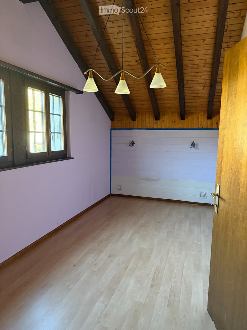 Empty room with wooden flooring, exposed wooden beams on the ceiling, and a hanging light fixture. The walls are painted in a light purple color, and there is a window on one side.