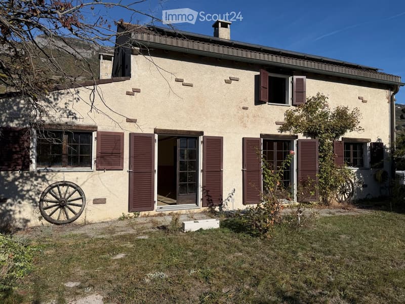 Two-story house with beige walls, red shutters, and a tiled roof. Features include a front door with glass panels, multiple windows, and a small garden area with grass and a few plants.
