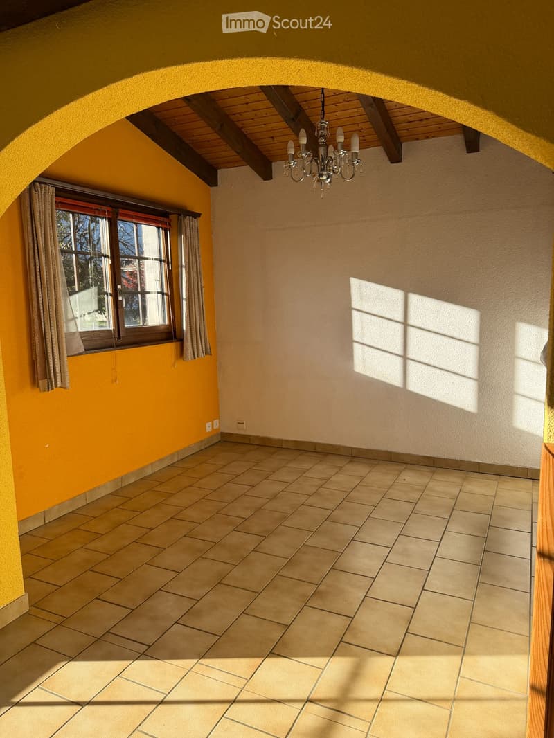Empty room with beige tiled floor, yellow and beige walls, large window with curtains, and a chandelier hanging from the ceiling.