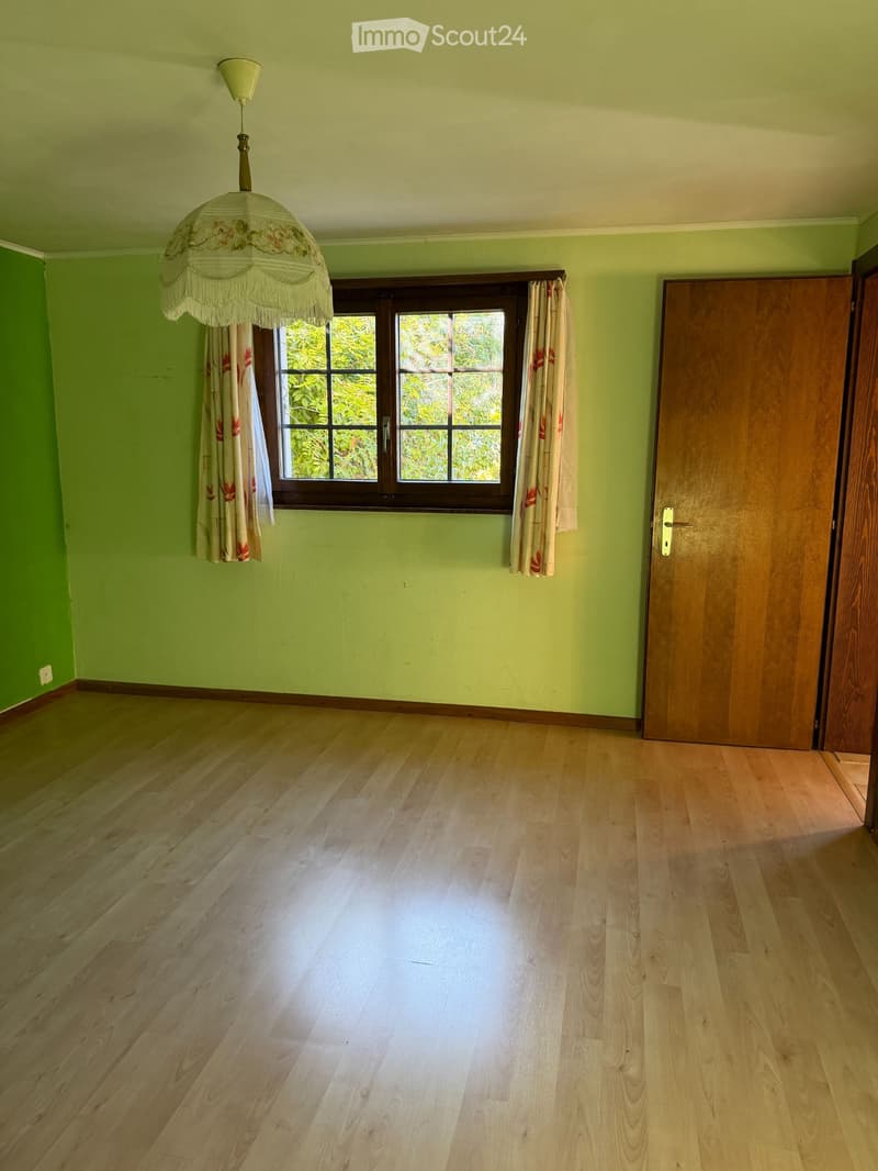 Empty room with light green walls, wooden floor, large window with curtains, and a wooden door.