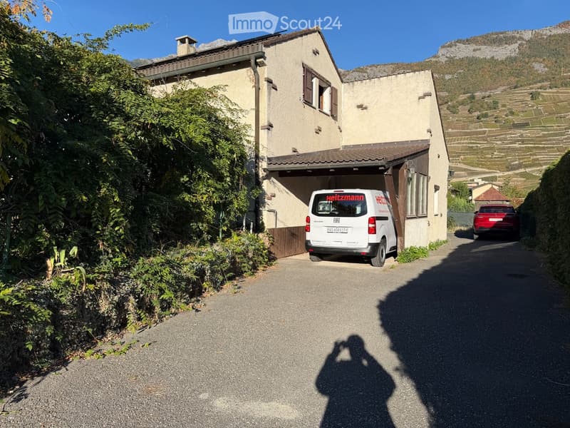 Two-story house with beige walls, wooden shutters, and a garage. A white van is parked in the driveway, with a red car visible further down the road. The house is surrounded by greenery and overlooks a vineyard and mountainous landscape.