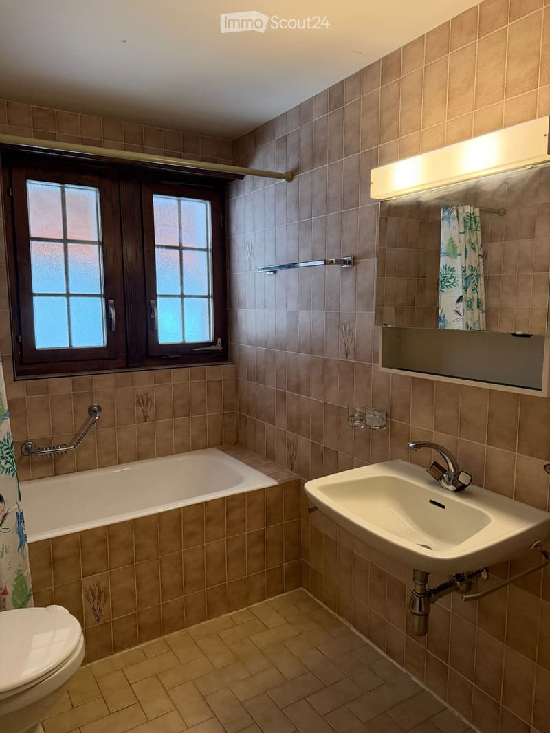 Bathroom with beige tiled walls and floor, featuring a bathtub, sink, toilet, mirror, and window with frosted glass panes.